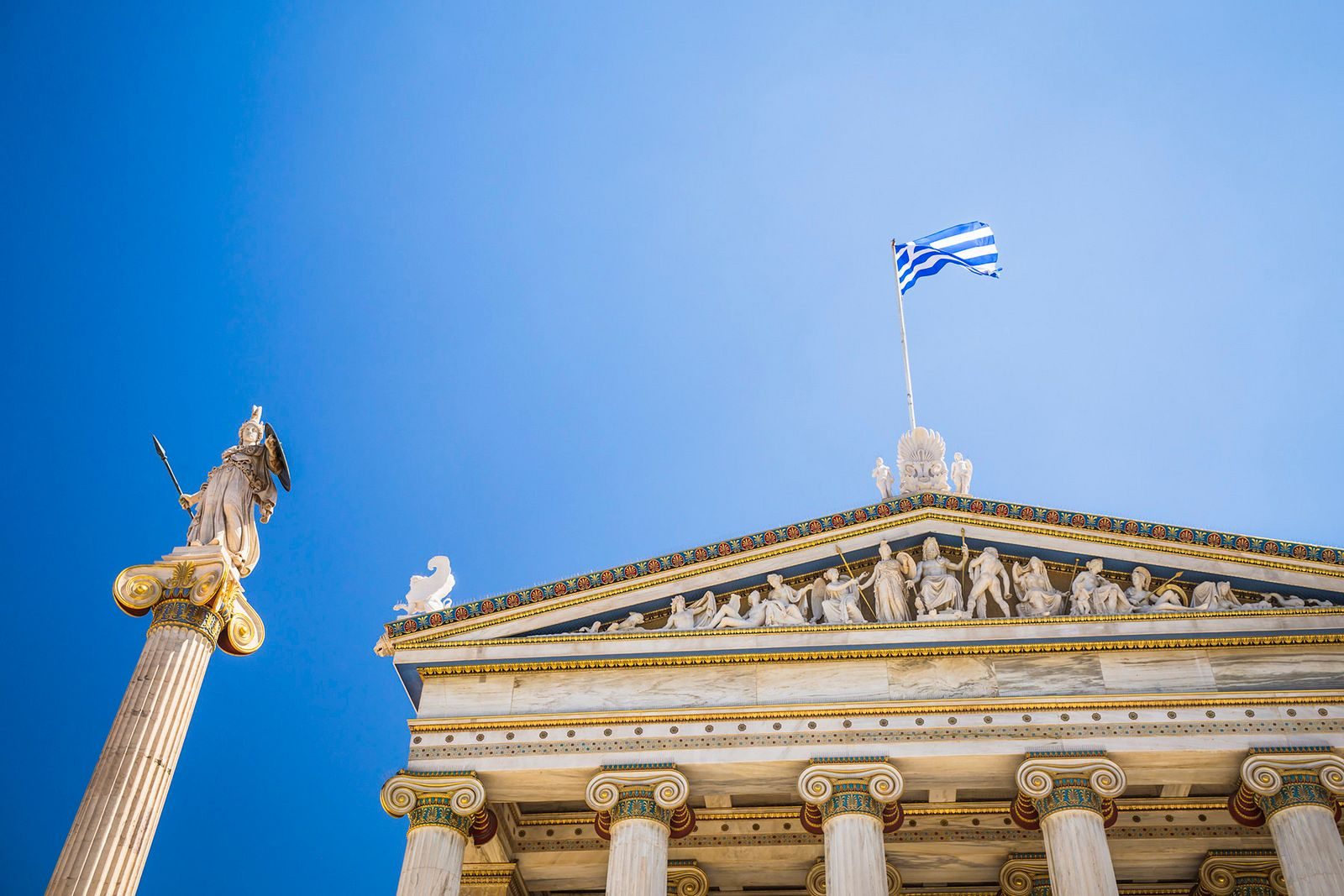 The Academy of Athens with a statue of Athena and the Greek flag against a blue sky