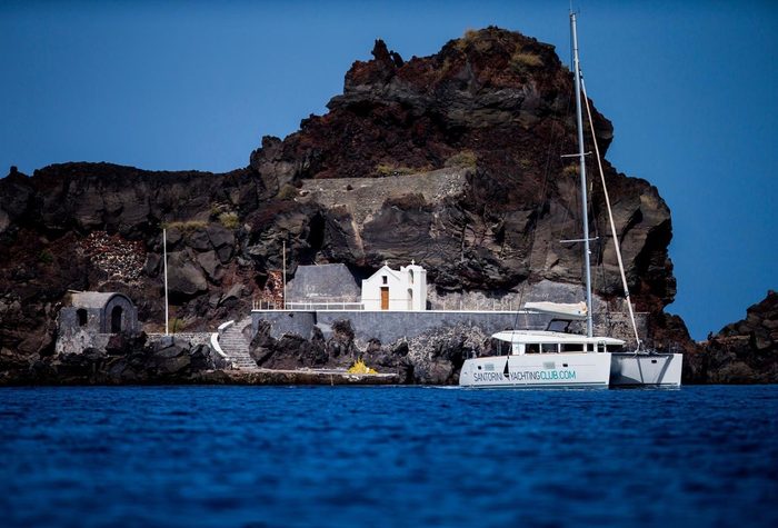 Catamaran moored beside a small white chapel at the foot of dark volcanic rock