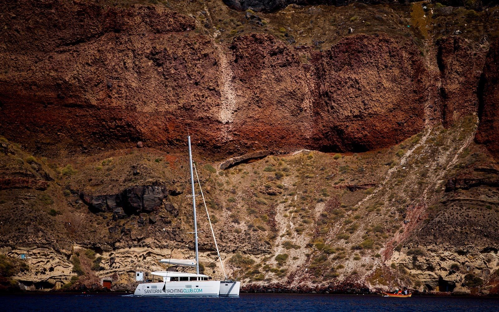 Sailing catamaran anchored below towering red and brown caldera cliffs