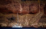 Sailing catamaran anchored below towering red and brown caldera cliffs