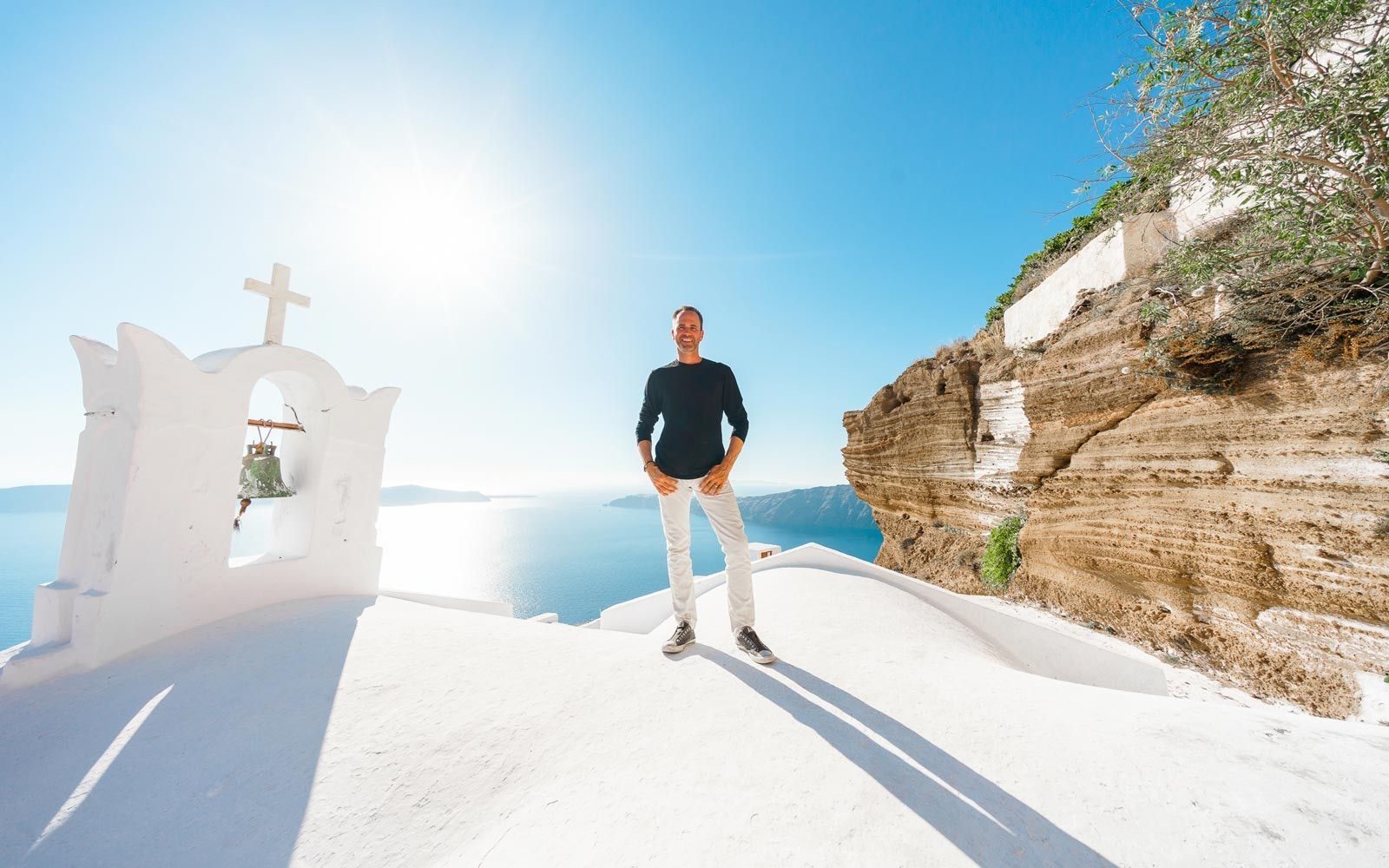 Man standing on a whitewashed church roof beside a bell gable, with the caldera behind