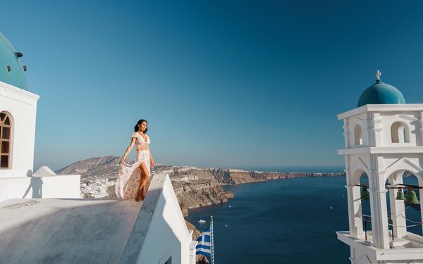 Woman in a floral dress on a church roof between a blue dome and a bell tower, caldera below