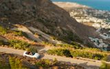 White convertible on a winding mountain road, with Kamari village and the sea below