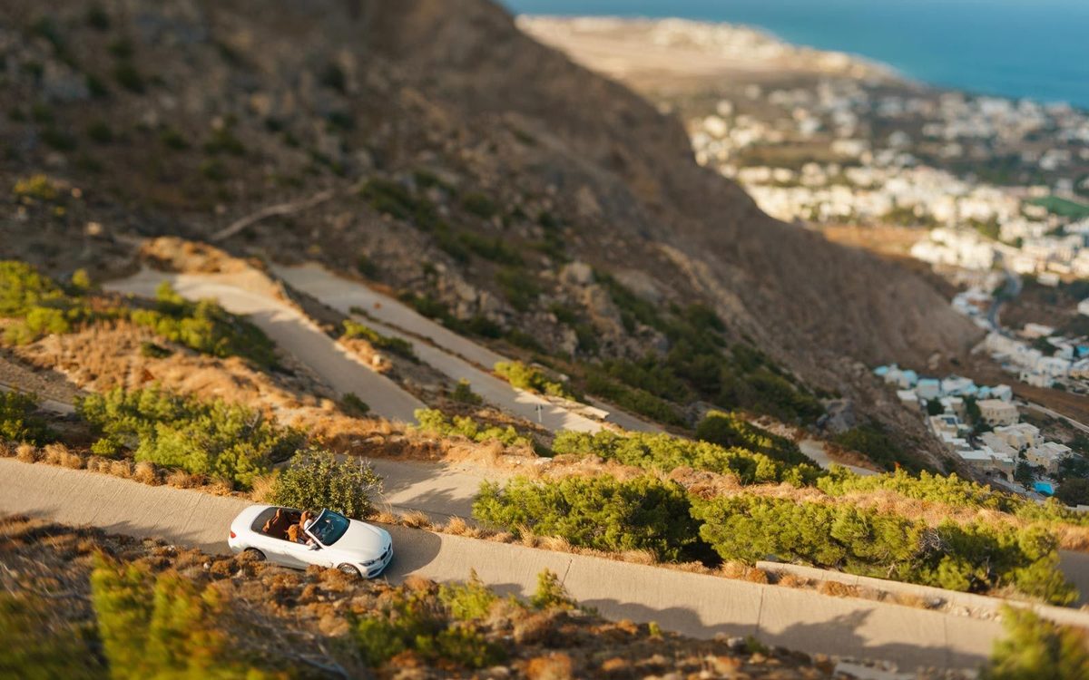 White convertible on a winding mountain road, with Kamari village and the sea below