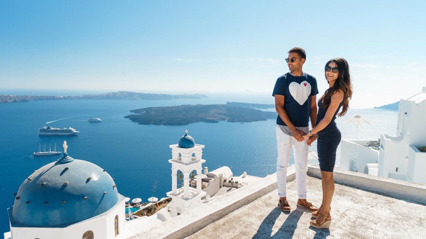 Couple in sunglasses on a caldera-view rooftop, with a blue dome and bell tower below