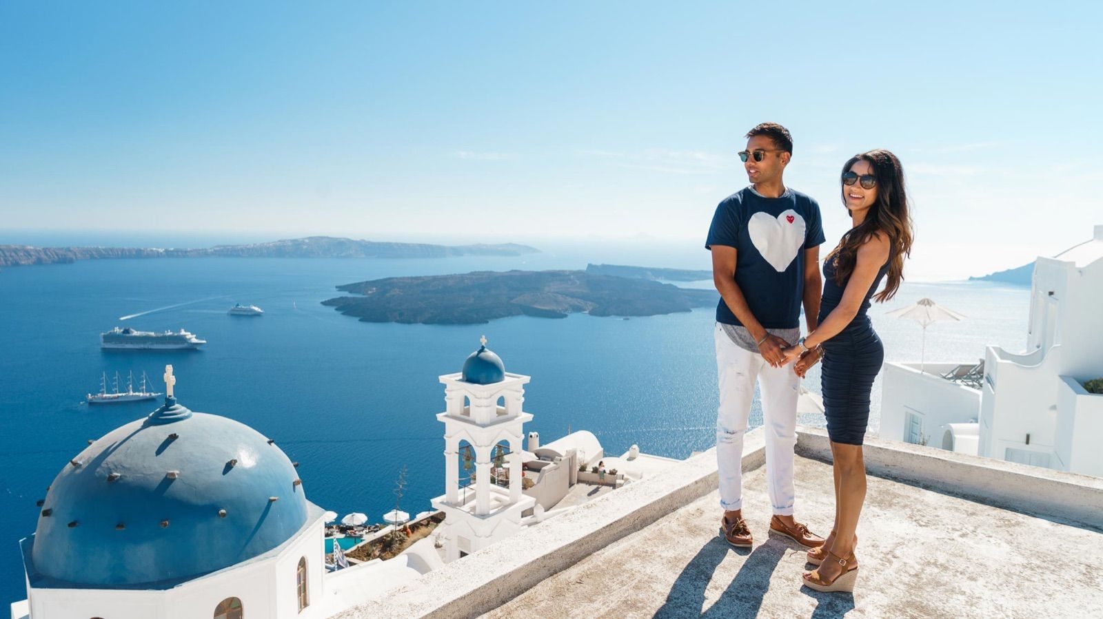 Couple in sunglasses on a caldera-view rooftop, with a blue dome and bell tower below