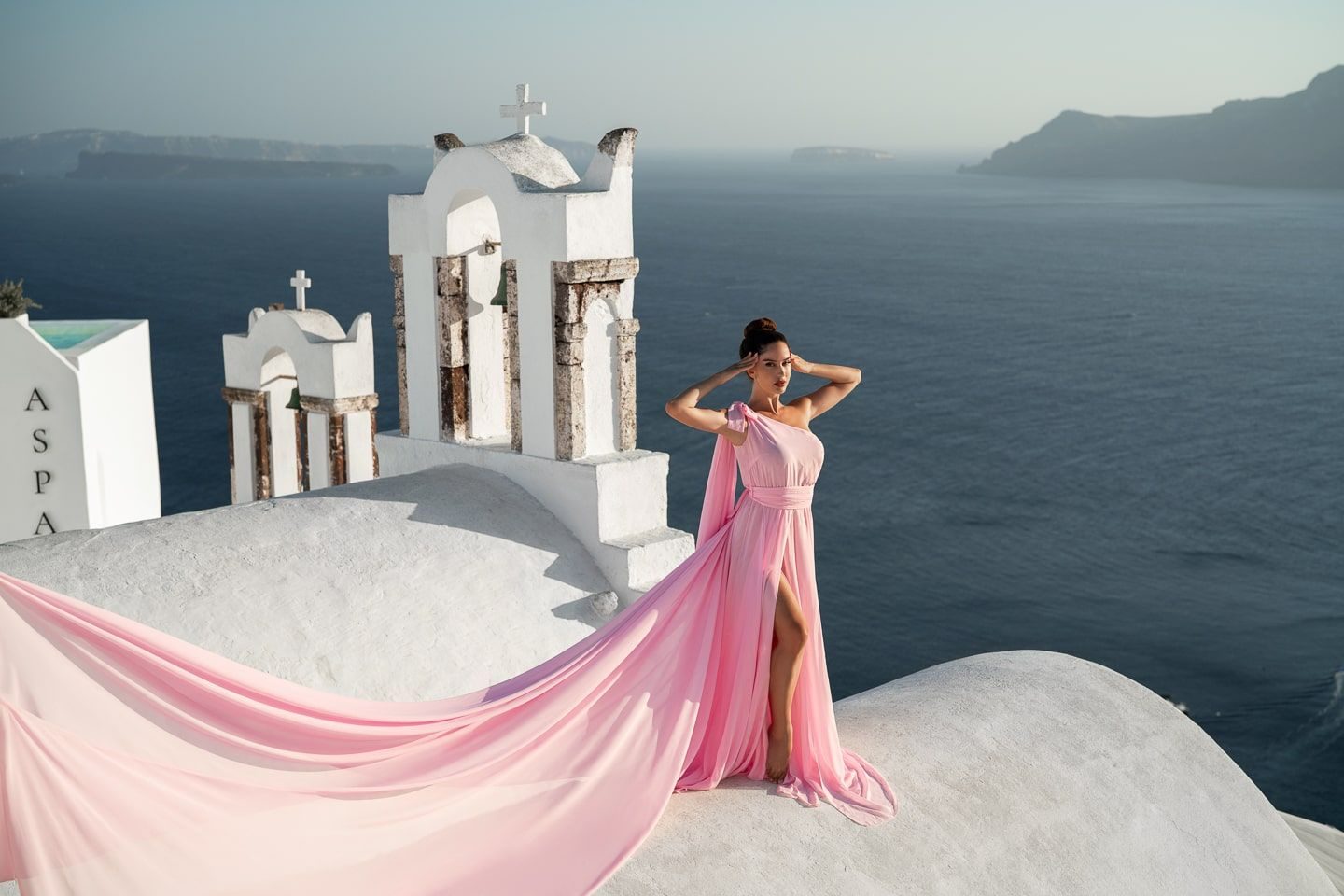 Woman in a long pink flying dress on a whitewashed rooftop beside a bell tower, above the Santorini caldera
