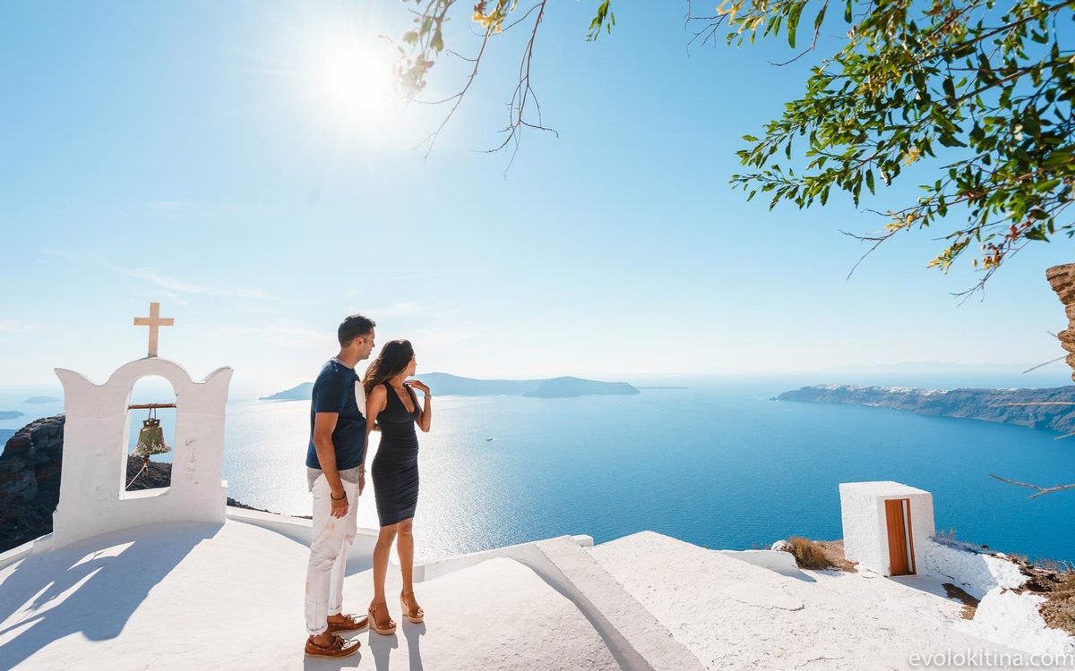 Couple on a whitewashed church roof beside a bell gable, overlooking the caldera