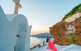 Couple by a white chapel in Imerovigli at sunset, the woman in a long pink dress