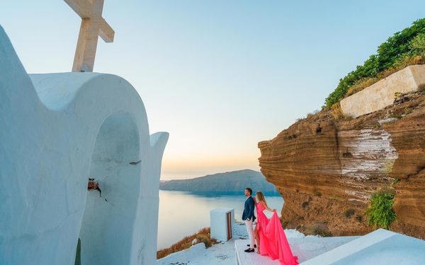 Couple by a white chapel in Imerovigli at sunset, the woman in a long pink dress