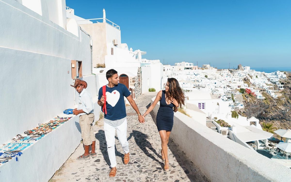 Couple walking hand in hand along a cobbled lane in Imerovigli, past a street seller