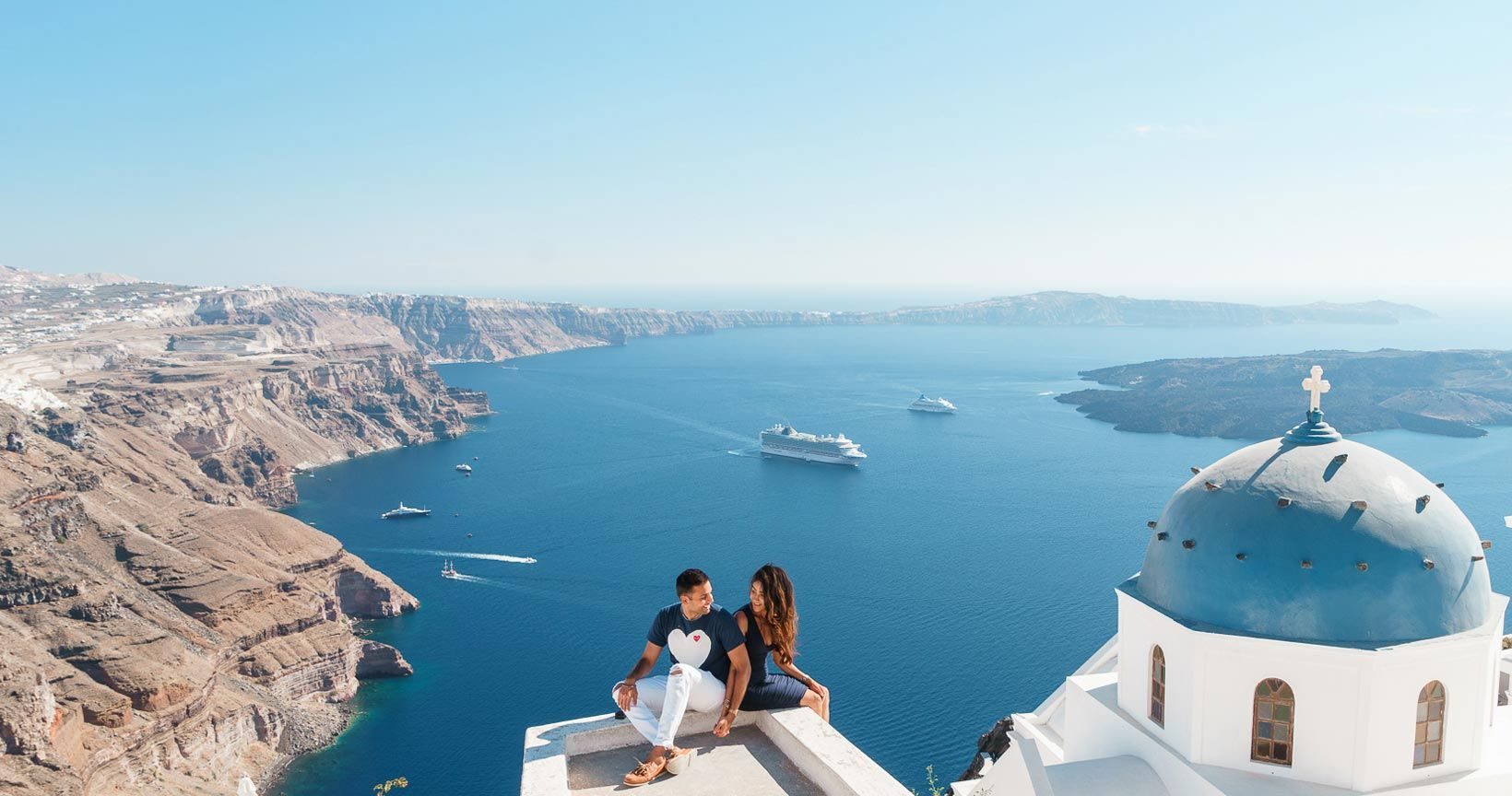 Couple on a rooftop in Imerovigli beside a blue dome, with cruise ships anchored in the caldera below