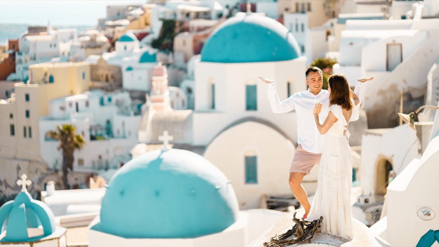 Couple in white with arms raised on a rooftop above the blue domes of Oia