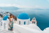 Woman in a floral skirt seated by the blue domes and pink bell tower of Oia