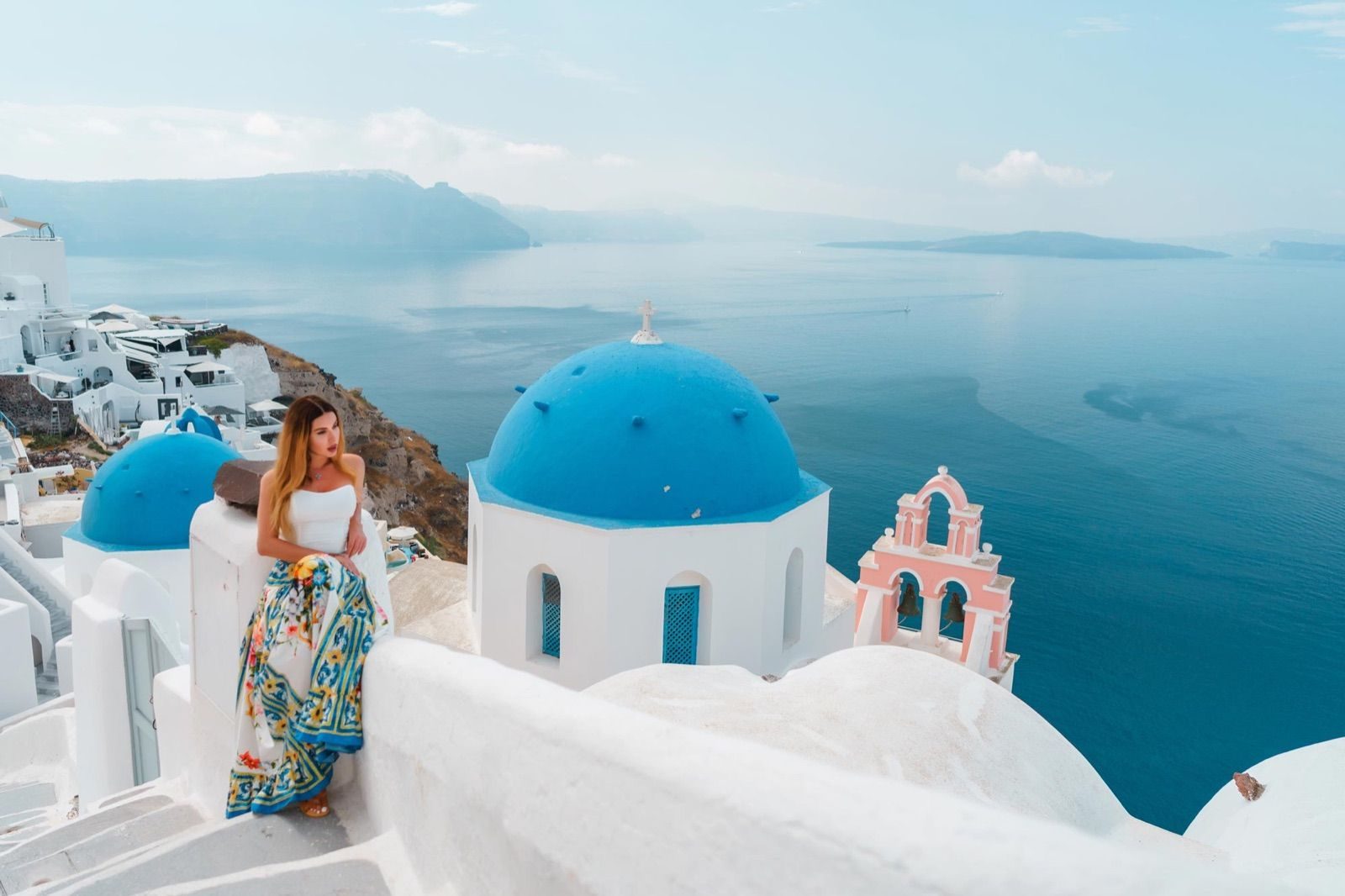 Woman in a floral skirt seated by the blue domes and pink bell tower of Oia