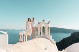 Couple jumping on a white church roof in Oia beside bell towers, caldera behind