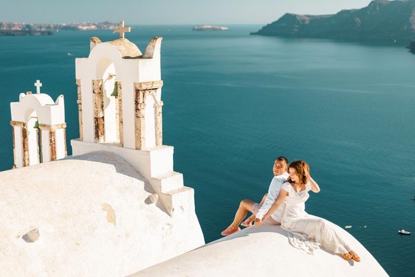 Couple sitting on a white church roof in Oia beside bell towers, looking over the caldera