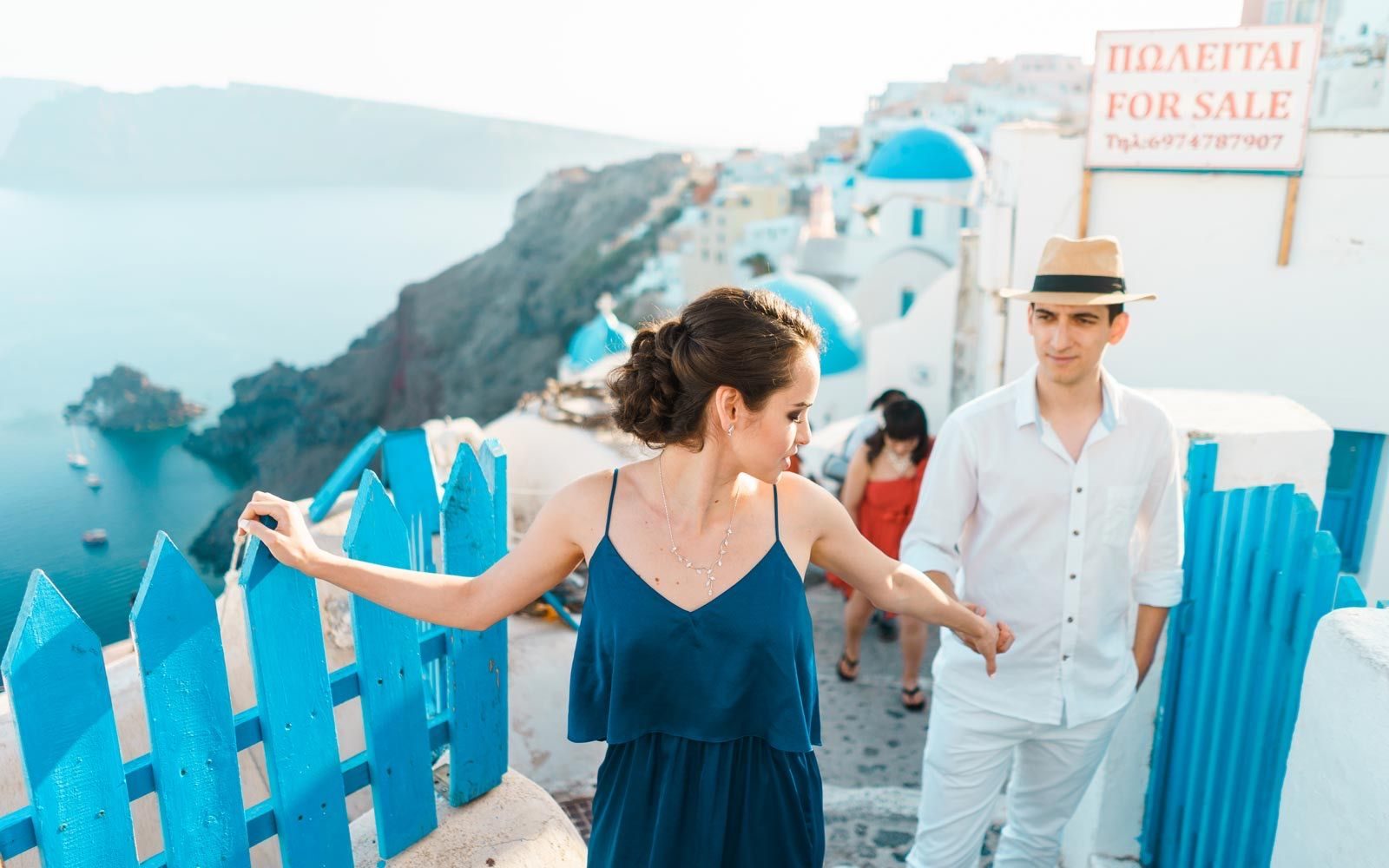 Couple on a lane in Oia by blue wooden gates, with blue domes and the caldera behind