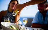 Olive oil poured over a Greek salad as two guests with white wine look on