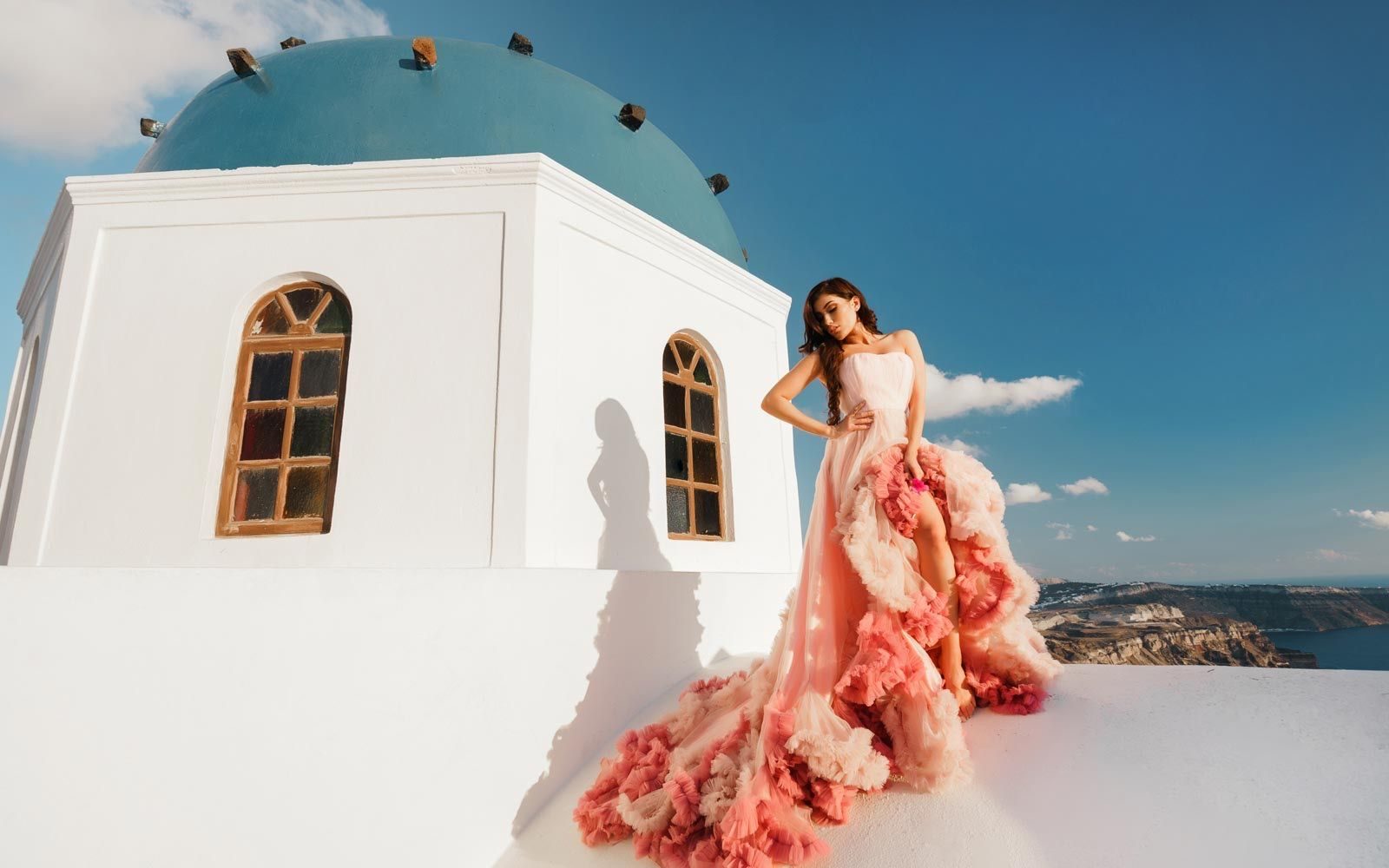 Woman in a pink ruffled gown beside a blue-domed church above the caldera