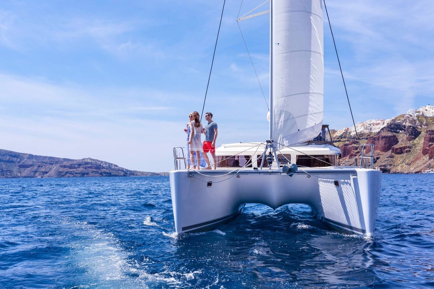 Couple at the bow of a sailing catamaran below red caldera cliffs and a clifftop village