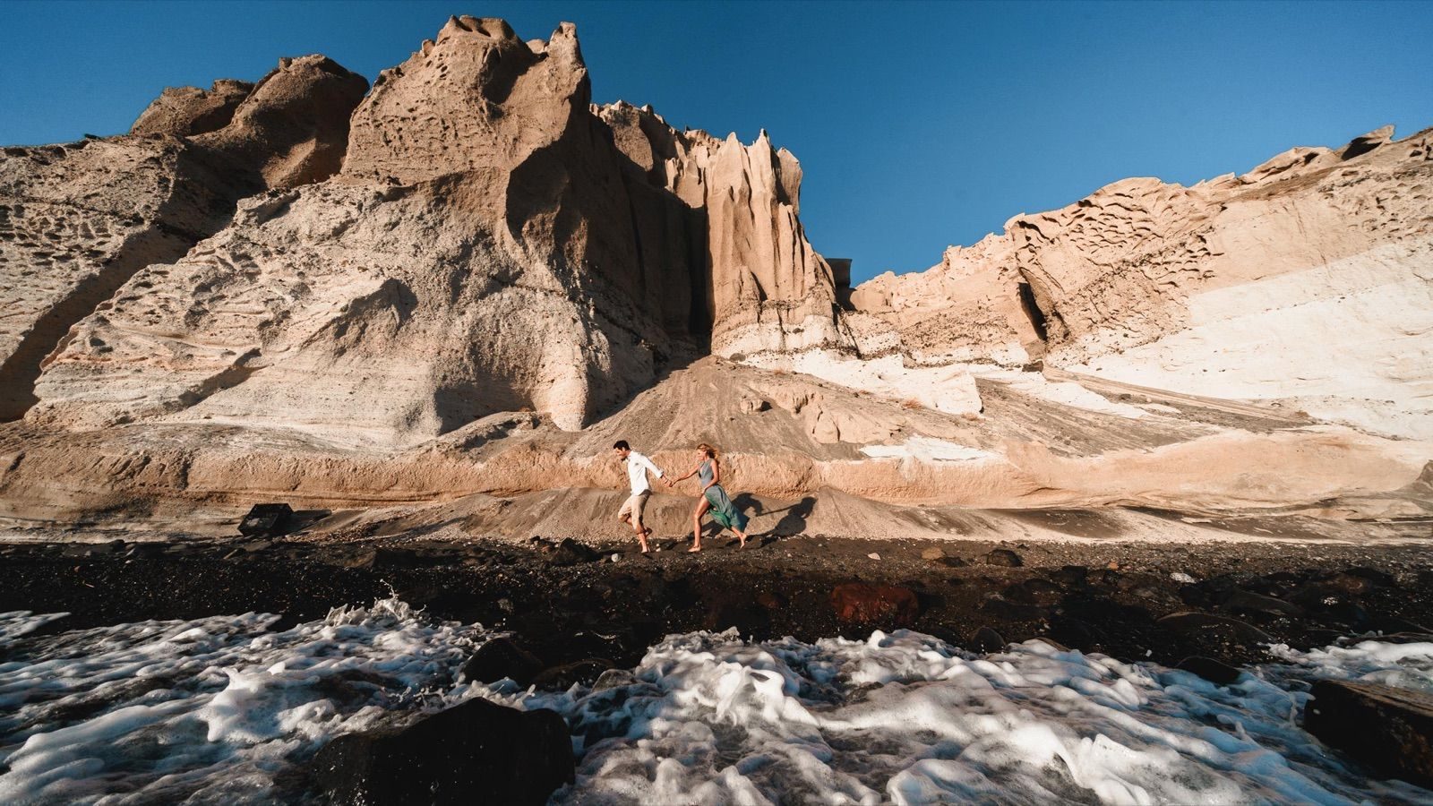 Couple hand in hand on a black volcanic beach, surf in front and eroded white cliffs behind