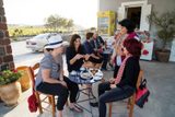 Guests sharing coffee and sweets at a café table outside a village shop, vineyards behind