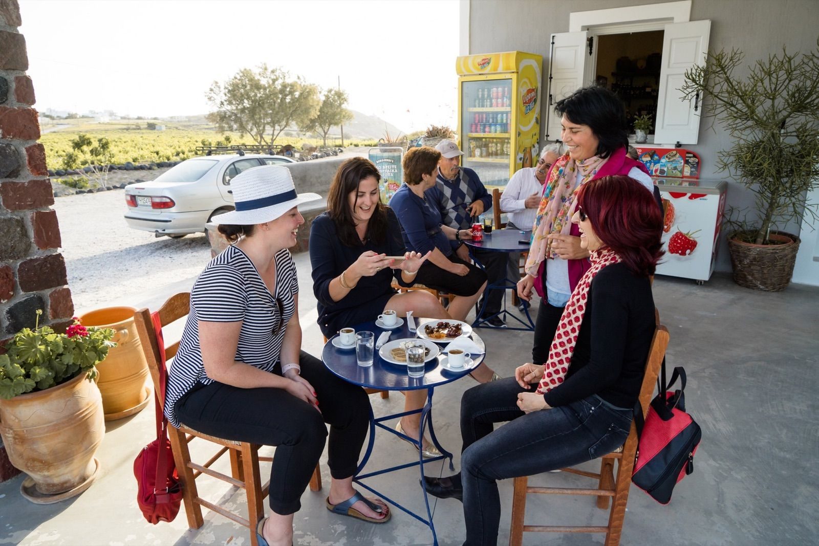 Guests sharing coffee and sweets at a café table outside a village shop, vineyards behind